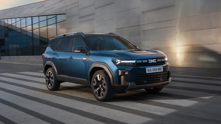 Plavi SUV vozi preko pješačkog prijelaza u urbanom okruženju, uz modernu zgradu sa staklenom fasadom u pozadini.