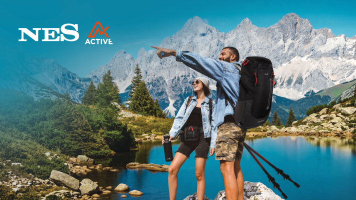Dvoje planinara s ruksacima stoje uz planinsko jezero i gledaju prema vrhovima planina, dok muškarac pokazuje u daljinu; u kutu je logo NES Active.