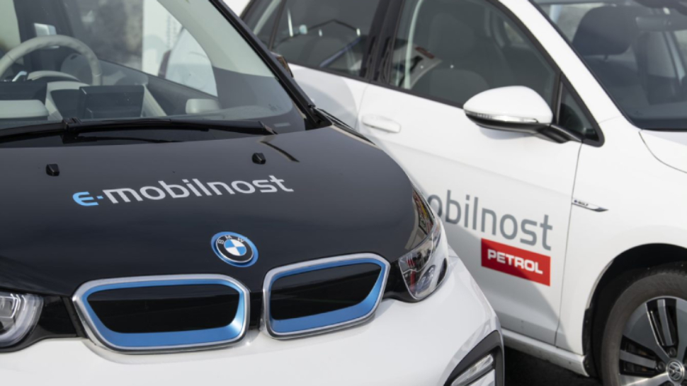 Na slici su dva električna automobila parkirana jedan pored drugoga. Na poklopcu motora i bočnim vratima vidljiv je natpis »e-mobilnost Petrol«. U prvom planu prikazan je prednji dio bijelog automobila BMW, dok je u pozadini vidljiv drugi automobil s Petrol oznakama.