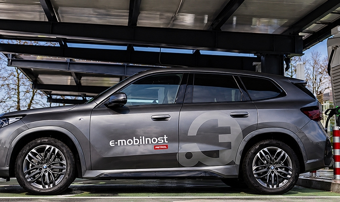 Na slici je sivi osobni automobil s natpisom »e-mobilnost Petrol«, parkiran na mjestu za punjenje električnih vozila. U pozadini su vidljivi nadstrešnica sa solarnim panelima, punionica za električna vozila i priključeni kabel za punjenje.