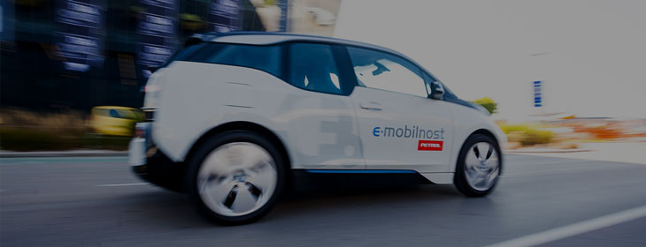 Na slici je bijeli električni automobil s natpisom »e-mobilnost Petrol« u vožnji gradskom prometnicom. Vozilo je prikazano iz bočne perspektive, a pozadina je zamućena zbog kretanja automobila.