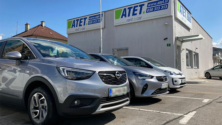 Na slici su automobili parkirani ispred poslovne zgrade s natpisom »ATET rent a car«. U prvom planu nalazi se sivi automobil, dok su iza njega parkirana još dva vozila. U pozadini su vidljivi ulaz u zgradu, parkiralište i reklamni natpisi na pročelju objekta.