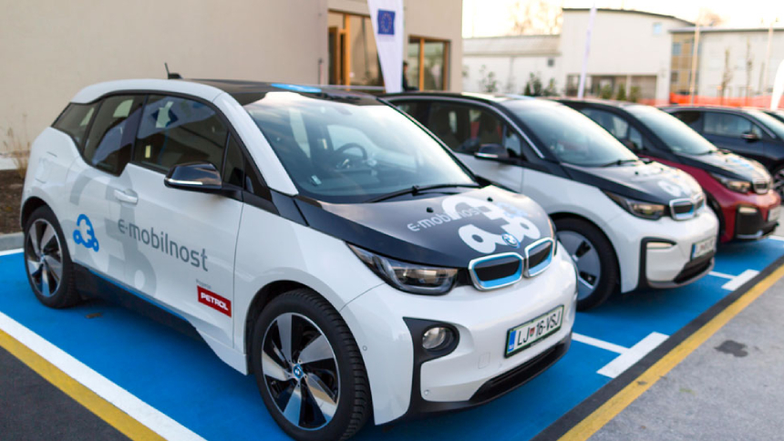 Na slici su tri električna automobila s oznakom »e-mobilnost Petrol« parkirana na označenim parkirnim mjestima. Vozila su bijele i crvene boje, a u pozadini su vidljivi poslovna zgrada, zastave i parkiralište.