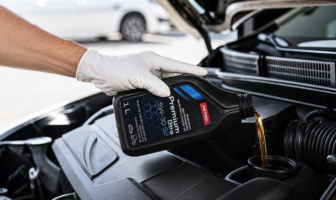 Ruka zaštićena bijelom rukavicom ulijeva motorno ulje Petrol Premium SW-30 S2 iz crne boce u motor automobila. Poklopac motora je otvoren, a u prvom planu vidljiv je postupak nadopunjavanja motornog ulja tijekom održavanja vozila. Slika simbolizira brigu o motoru, redovito servisiranje i optimalne performanse vozila.