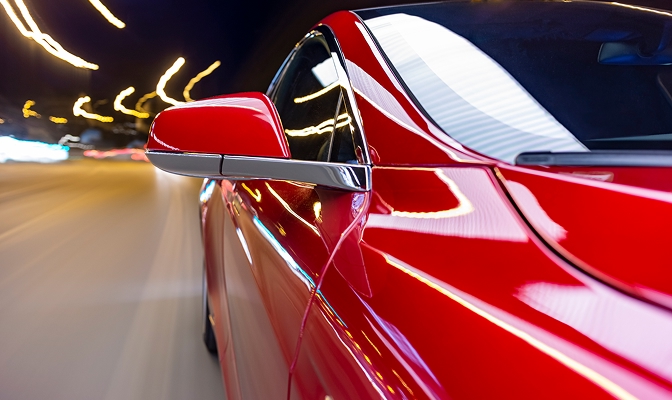 Na slici je prikazan detaljan bočni pogled na crveni osobni automobil u vožnji. U prvom planu istaknuto je vanjsko bočno ogledalo i sjajna karoserija vozila, dok su u pozadini vidljivi zamućeni tragovi svjetala koji stvaraju dojam brzine, kretanja i dinamične noćne vožnje kroz osvijetljeni gradski ili prometni prostor.
