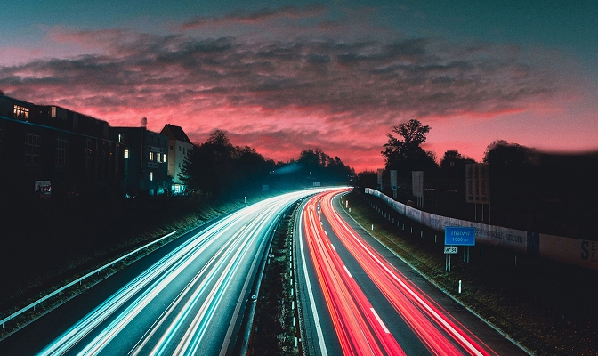 Noćni prizor autoceste s dugim svjetlosnim tragovima vozila u bijeloj i crvenoj boji. Cesta prolazi između zgrada i drveća, dok je nebo u pozadini obasjano ružičastim i crvenim tonovima zalaska sunca.