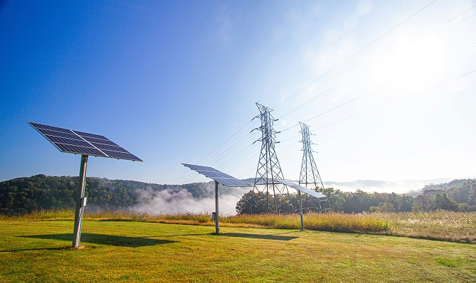 Na slici su solarni paneli postavljeni na travnatoj površini. U pozadini se nalaze visokonaponski dalekovodi i šumski krajolik. Nebo je vedro i plave boje, a sunčeva svjetlost osvjetljava cijeli prizor.