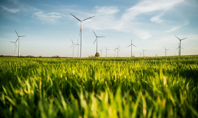 Na slici su vjetroturbine postavljene na travnatoj površini. U prvom planu vidi se zelena trava, dok su u pozadini raspoređene vjetroturbine različitih veličina. Iznad krajolika pruža se vedro nebo s rijetkim oblacima.