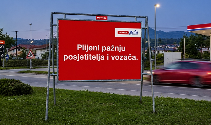 Veliki crveni reklamni pano uz prometnicu s porukom ‘Plijeni pažnju posjetitelja i vozača’, dok u pozadini prolazi automobil.