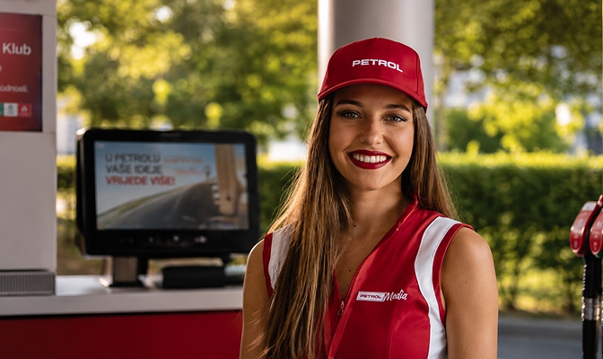 Nasmejana promotorka u crvenoj uniformi i kapi s logotipom Petrola stoji na prodajnom mjestu, uz digitalni zaslon u pozadini.