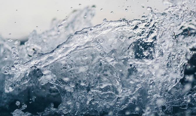 Na slici je krupni prikaz vode u pokretu. Vidljivi su valovi, mjehurići i kapljice vode različitih veličina, dok je pozadina zamućena i u svijetlim tonovima.